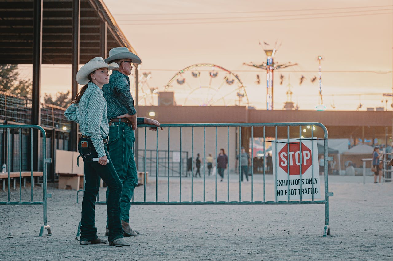 services-04 Two cowboys in hats lean on a railing, with a stop sign and fair rides in the background at sunset.