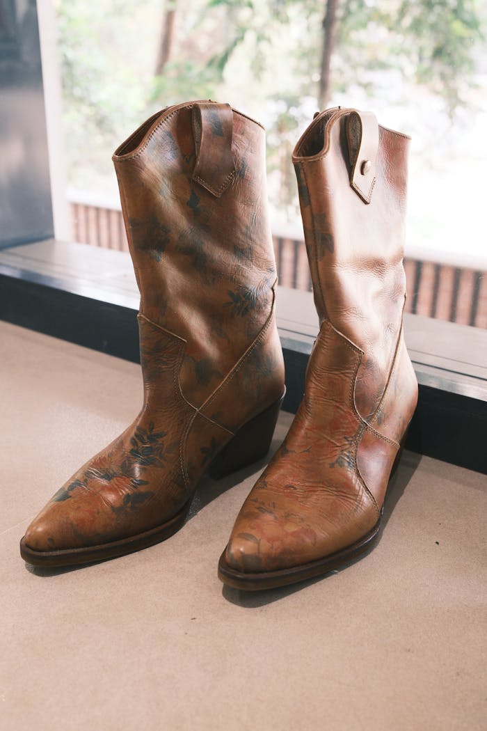 services-02 Pair of vintage leather cowboy boots with floral design placed indoors near a window.