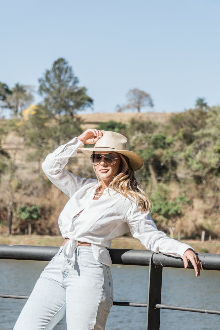 services-01 Chic woman in white blouse and hat posing by a scenic riverside railing on a sunny day.