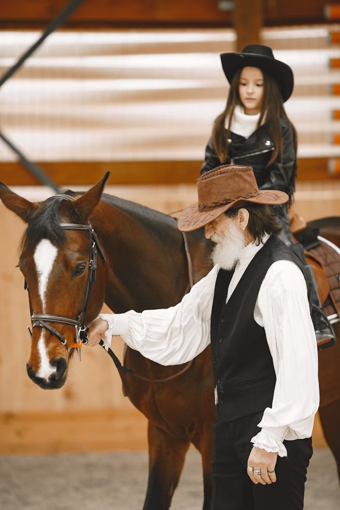 who-we-are An elderly cowboy assists a young cowgirl riding a brown horse indoors, showcasing equestrian skills.