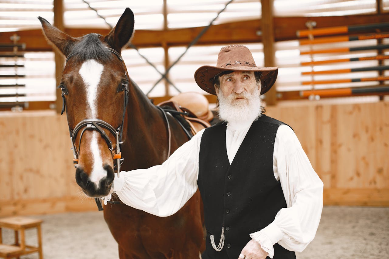 services-03 Elderly man in cowboy hat beside a brown horse, standing in a rustic barn.